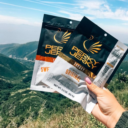 An arm in a blue jacket holds two packages of Perky Jerky, a caffeinated turkey jerky. The packs are black, white, and gold with the title and a stylized turkey logo..